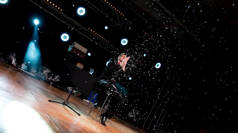 Iluzjonista Konrad Mościński podczas nowoczesnego pokazu iluzji na dużej scenie. Artysta siedzi na krześle i wyczarowuje śnieg na tle widowiskowego oświetlenia scenicznego.