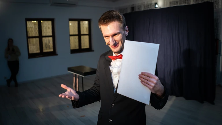Uśmiechnięty Iluzjonista Konrad Mościński przeprowadzający dialog z widzem. W ręce trzyma kopertę z przepowiednią.