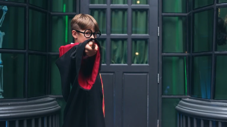 Młody chłopiec w okrągłych okularach i czarno-czerwonej szacie czarodzieja, przypominającej strój Harry'ego Pottera